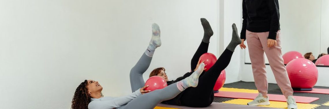 Two women exercising with pink balls on yoga mats under trainer supervision in a Yalova gym.